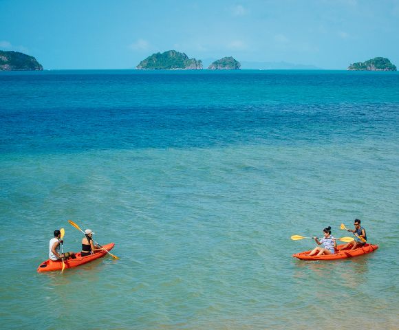 Two orange kayaks with people paddling on clear turquoise water, with small green islands in the distance under a sunny blue sky.