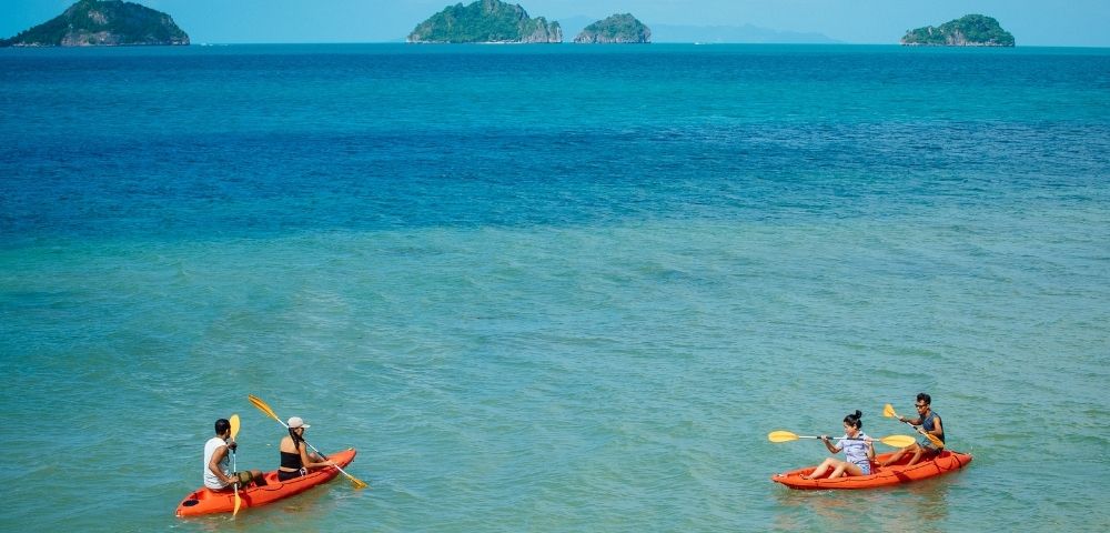 Two orange kayaks with people paddling on clear turquoise water, with small green islands in the distance under a sunny blue sky.