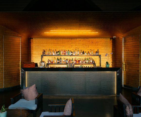 Sophisticated bar interior with dim lighting, warm golden hues, a stocked shelf of bottles, and cozy seating with plush pillows, creating an inviting ambiance.