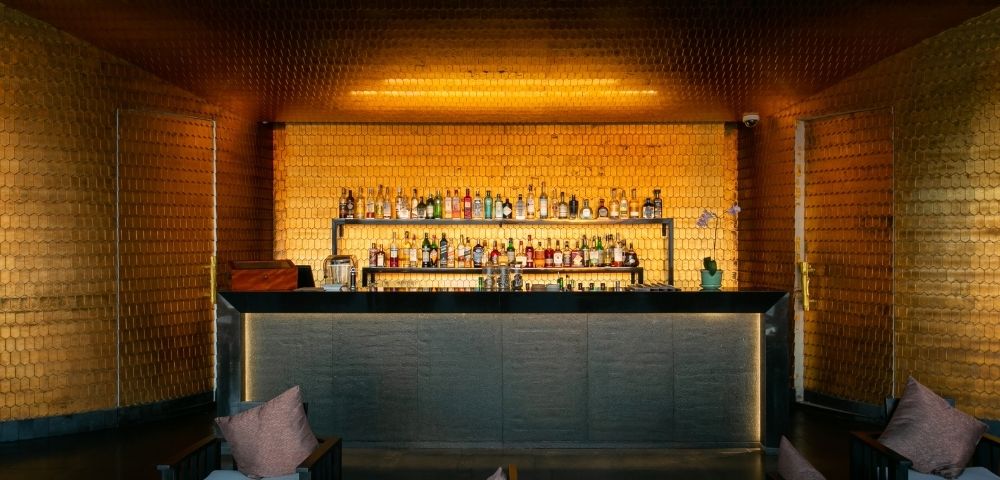 Sophisticated bar interior with dim lighting, warm golden hues, a stocked shelf of bottles, and cozy seating with plush pillows, creating an inviting ambiance.