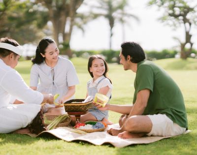 A family enjoys a picnic on a sunny day, sitting on a mat in a park. They are smiling and sharing food, surrounded by grass and trees.