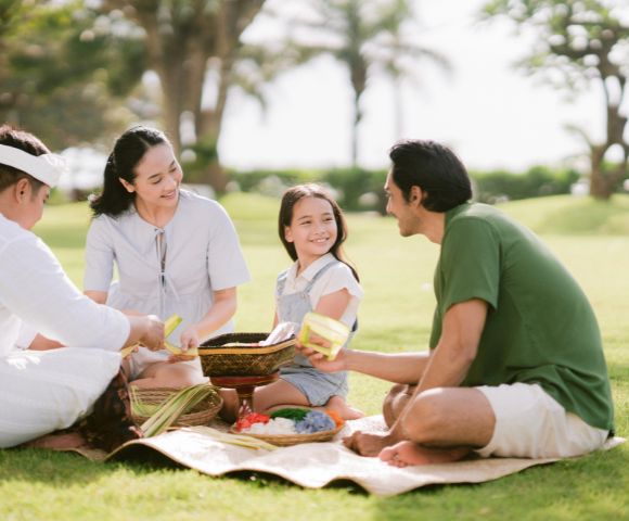 A family enjoys a picnic on a sunny day, sitting on a mat in a park. They are smiling and sharing food, surrounded by grass and trees.