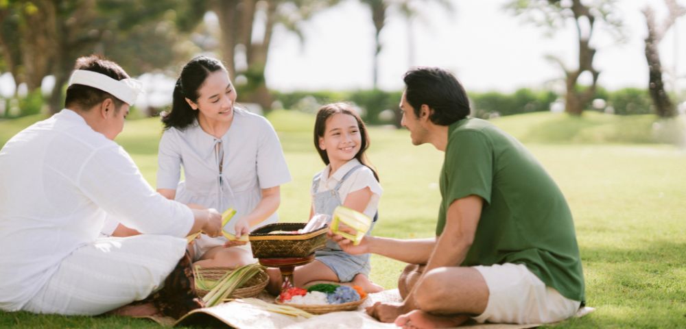 A family enjoys a picnic on a sunny day, sitting on a mat in a park. They are smiling and sharing food, surrounded by grass and trees.