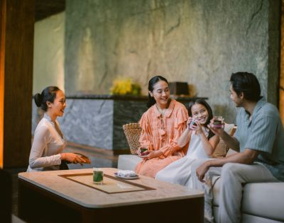 A family of four enjoys tea on a cozy sofa, smiling warmly in a serene, softly lit room with textured walls and a minimalist design.