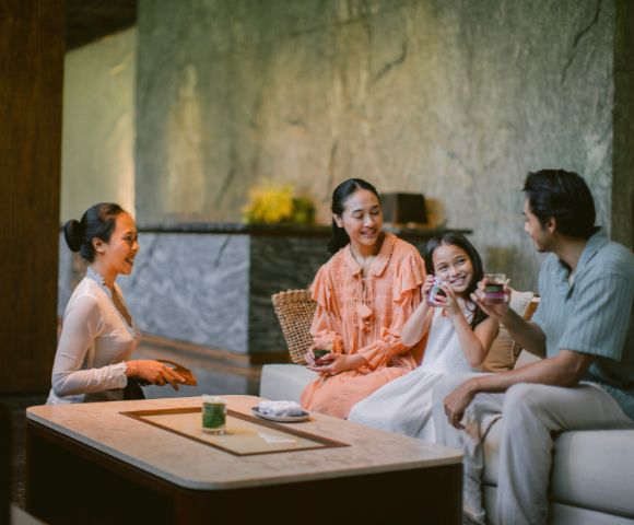 A family of four enjoys tea on a cozy sofa, smiling warmly in a serene, softly lit room with textured walls and a minimalist design.