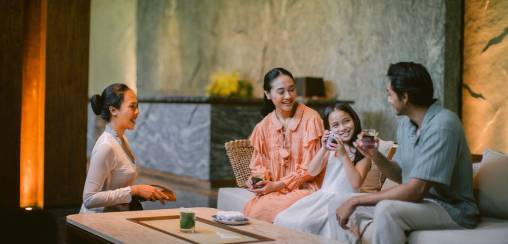 A family of four enjoys tea on a cozy sofa, smiling warmly in a serene, softly lit room with textured walls and a minimalist design.