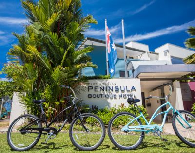 Two bicycles, one black and one blue, are parked on grass in front of the Peninsula Boutique Hotel. Palm trees and clear blue sky add to the tropical vibe.