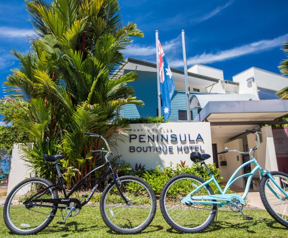 Two bicycles, one black and one blue, are parked on grass in front of the Peninsula Boutique Hotel. Palm trees and clear blue sky add to the tropical vibe.