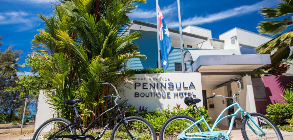 Two bicycles, one black and one blue, are parked on grass in front of the Peninsula Boutique Hotel. Palm trees and clear blue sky add to the tropical vibe.