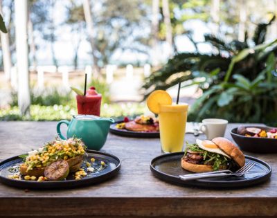 Outdoor brunch setting with a vibrant burger, avocado toast topped with corn, fresh juice, and coffee. Lush greenery and ocean view in the background.