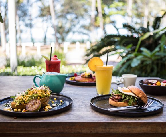 Outdoor brunch setting with a vibrant burger, avocado toast topped with corn, fresh juice, and coffee. Lush greenery and ocean view in the background.