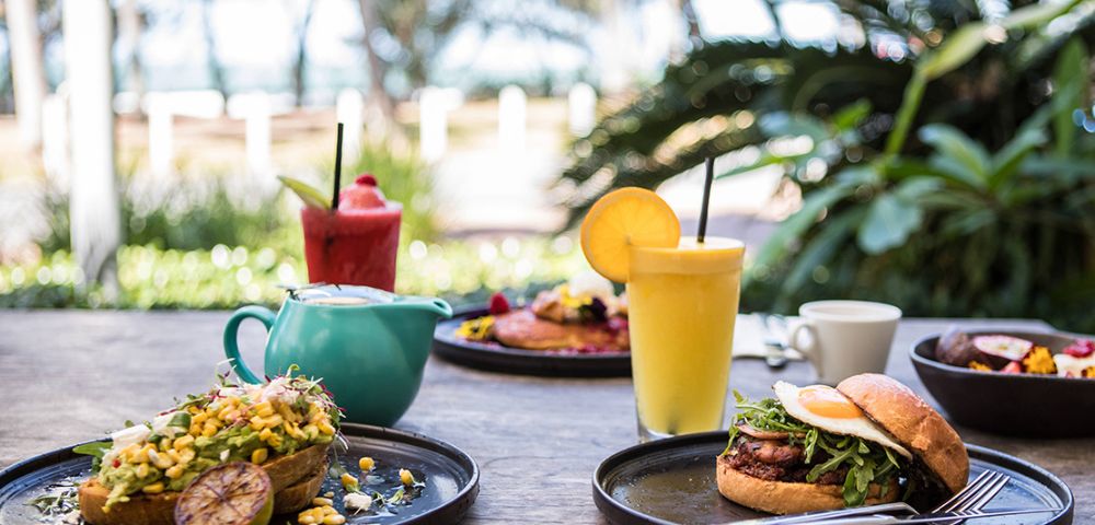 Outdoor brunch setting with a vibrant burger, avocado toast topped with corn, fresh juice, and coffee. Lush greenery and ocean view in the background.