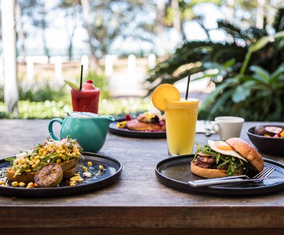 A vibrant outdoor brunch setting on a wooden table featuring an avocado dish, burger, smoothies, and a teapot. Lush greenery in the background.