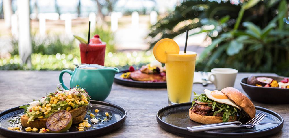 A vibrant outdoor brunch setting on a wooden table featuring an avocado dish, burger, smoothies, and a teapot. Lush greenery in the background.