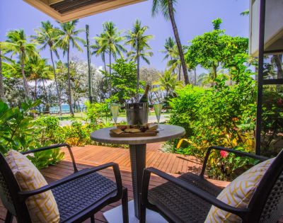 Two wicker chairs with yellow cushions face a small table holding a bucket with drinks, set on a wooden deck surrounded by lush greenery and palm trees.