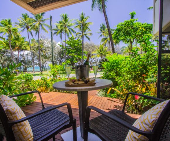 Two wicker chairs with yellow cushions face a small table holding a bucket with drinks, set on a wooden deck surrounded by lush greenery and palm trees.