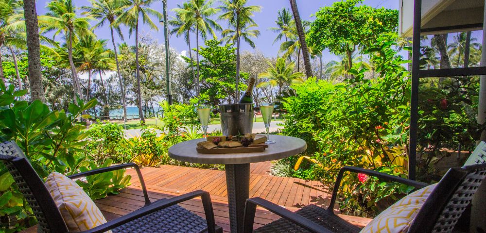Two wicker chairs with yellow cushions face a small table holding a bucket with drinks, set on a wooden deck surrounded by lush greenery and palm trees.