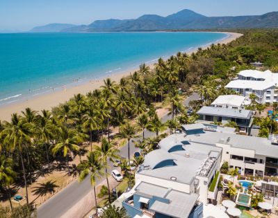 Aerial view of a tropical beach with turquoise ocean, lined by palm trees and white buildings. Mountains are visible in the distance under a clear blue sky.