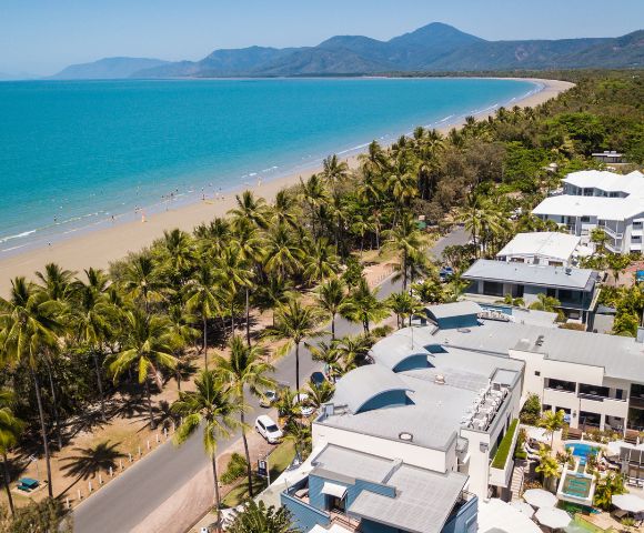 Aerial view of a tropical beach with turquoise ocean, lined by palm trees and white buildings. Mountains are visible in the distance under a clear blue sky.