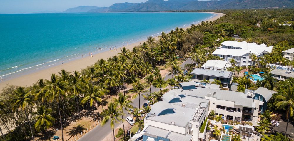 Aerial view of a tropical beach with turquoise ocean, lined by palm trees and white buildings. Mountains are visible in the distance under a clear blue sky.