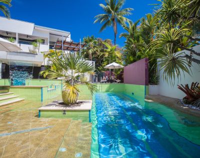 Luxurious poolside view with turquoise water and surrounding palm trees. Modern white building in the background, under a vibrant blue sky.