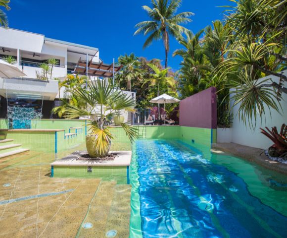 Luxurious poolside view with turquoise water and surrounding palm trees. Modern white building in the background, under a vibrant blue sky.