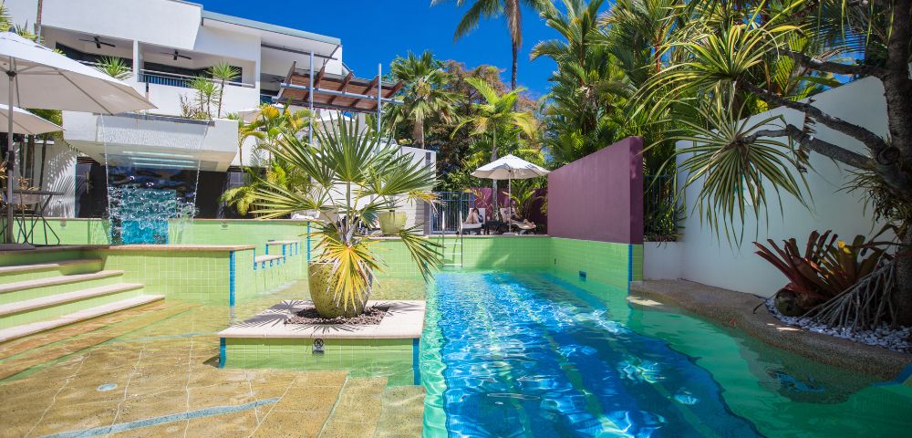 Luxurious poolside view with turquoise water and surrounding palm trees. Modern white building in the background, under a vibrant blue sky.
