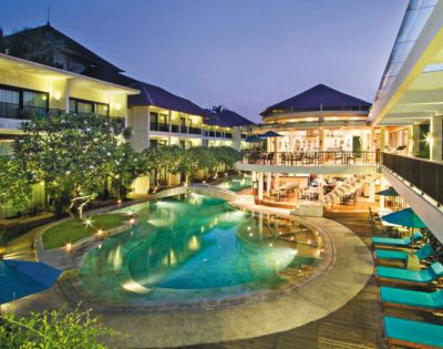 Luxurious hotel with a central, glowing pool surrounded by lush greenery at dusk. Elegant buildings and cozy poolside seating create a serene ambiance.