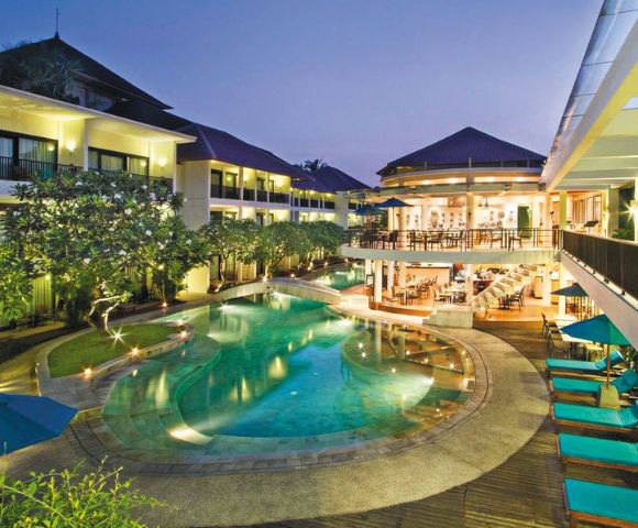 Luxurious hotel with a central, glowing pool surrounded by lush greenery at dusk. Elegant buildings and cozy poolside seating create a serene ambiance.