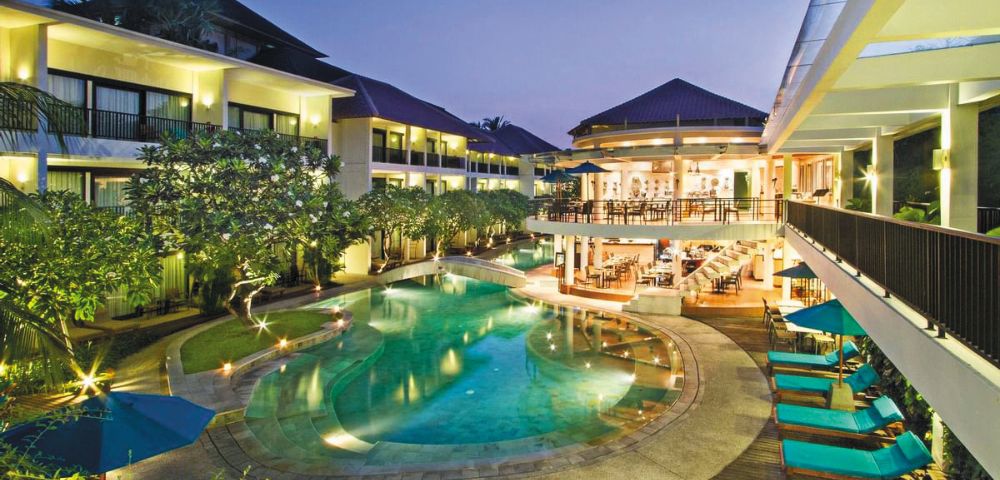 Luxurious hotel with a central, glowing pool surrounded by lush greenery at dusk. Elegant buildings and cozy poolside seating create a serene ambiance.
