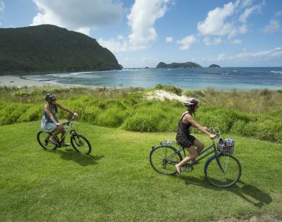 Two people ride bicycles on a grassy hill overlooking a calm ocean and distant islands under a blue sky, conveying a sense of freedom and tranquility.