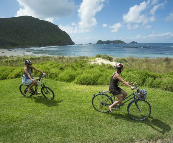 Two people ride bicycles on a grassy hill overlooking a calm ocean and distant islands under a blue sky, conveying a sense of freedom and tranquility.