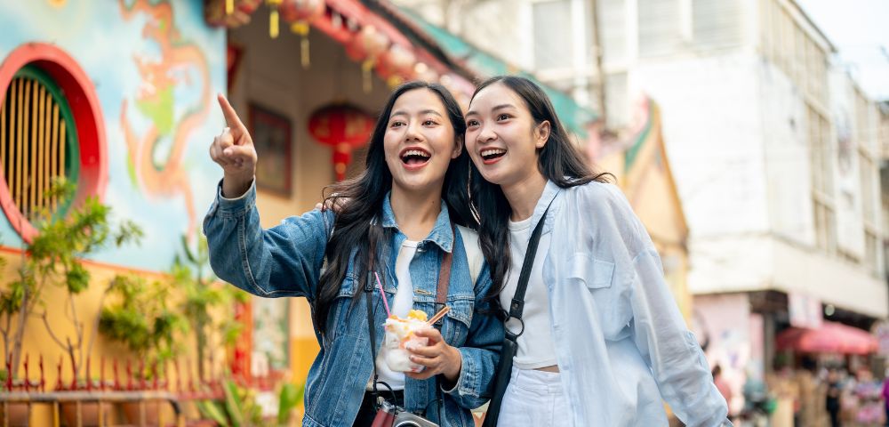 Two smiling women, one pointing, enjoy a vibrant street scene with colorful architecture in the background. They radiate joy and excitement.