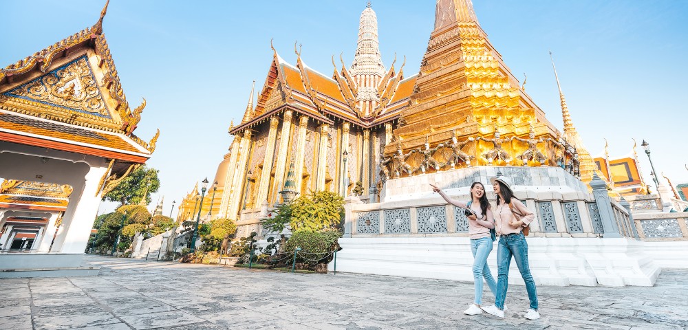Two people joyfully pose in front of a dazzling golden temple with intricate architecture under a clear blue sky, conveying a sense of wonder and happiness.