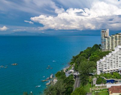 Coastal view with high-rise buildings, lush green trees, and turquoise ocean under a sky filled with dramatic clouds. Tranquil and scenic atmosphere.
