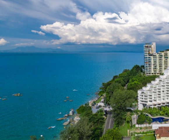 Coastal view with high-rise buildings, lush green trees, and turquoise ocean under a sky filled with dramatic clouds. Tranquil and scenic atmosphere.