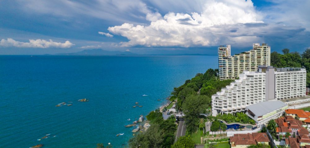 Coastal view with high-rise buildings, lush green trees, and turquoise ocean under a sky filled with dramatic clouds. Tranquil and scenic atmosphere.