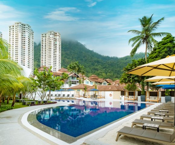 A serene poolside scene with lounge chairs and umbrellas, surrounded by palm trees. High-rise buildings and a lush green hill under a bright blue sky in the background.