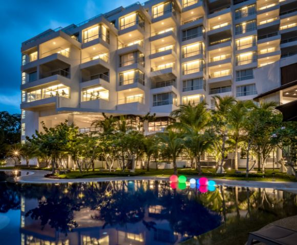 Illuminated hotel building at night with warm lights, a reflective pool in the foreground, surrounded by palm trees, and glowing colorful spheres.