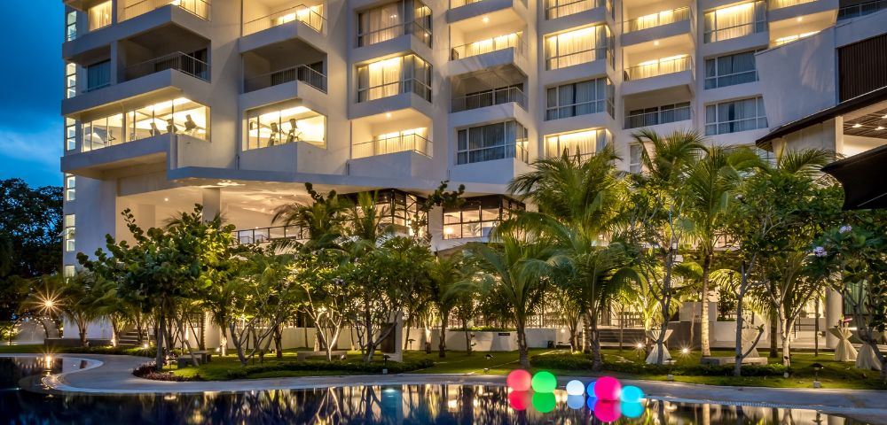 Illuminated hotel building at night with warm lights, a reflective pool in the foreground, surrounded by palm trees, and glowing colorful spheres.