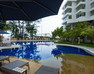 Modern hotel poolside with clear blue water, surrounded by loungers and umbrellas. Nearby palm trees and a white building create a tranquil, inviting atmosphere.