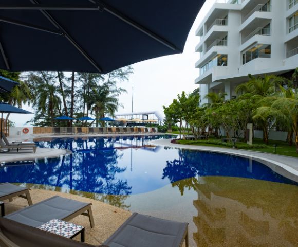 Modern hotel poolside with clear blue water, surrounded by loungers and umbrellas. Nearby palm trees and a white building create a tranquil, inviting atmosphere.