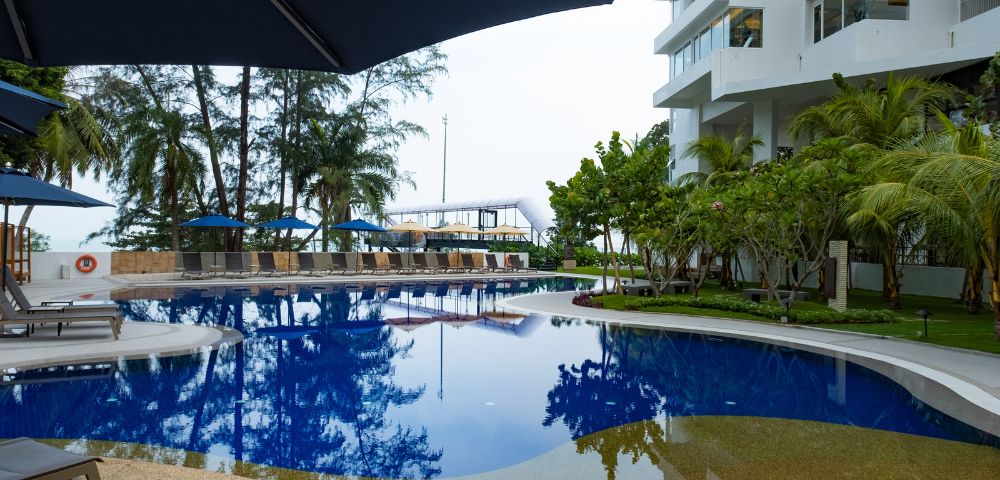 Modern hotel poolside with clear blue water, surrounded by loungers and umbrellas. Nearby palm trees and a white building create a tranquil, inviting atmosphere.