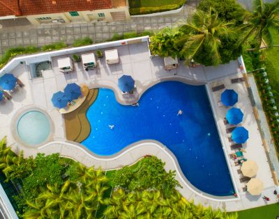 Aerial view of a vibrant blue pool with sunbeds and umbrellas. The pool has a circular kiddie area and tropical greenery surrounds the deck.