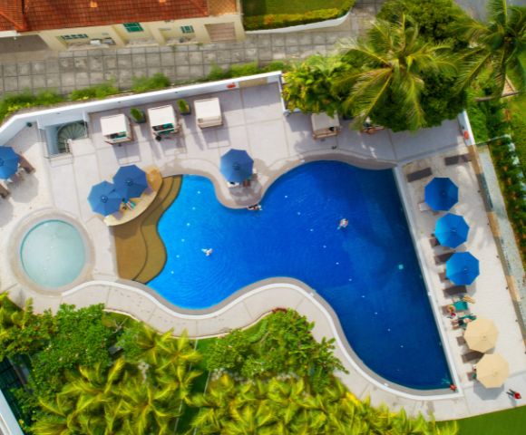 Aerial view of a vibrant blue pool with sunbeds and umbrellas. The pool has a circular kiddie area and tropical greenery surrounds the deck.