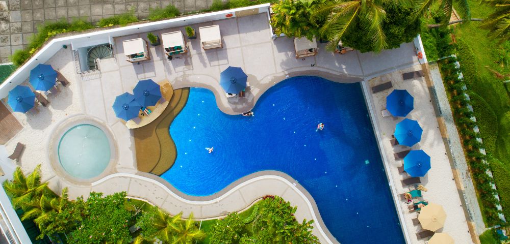 Aerial view of a vibrant blue pool with sunbeds and umbrellas. The pool has a circular kiddie area and tropical greenery surrounds the deck.