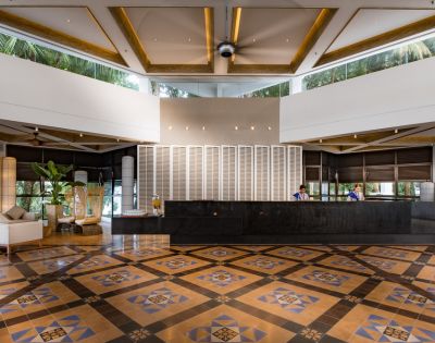 Spacious hotel lobby with a geometric patterned floor, dark reception desk, and modern decor. Two staff members are behind the desk, and lush greenery is visible outside.