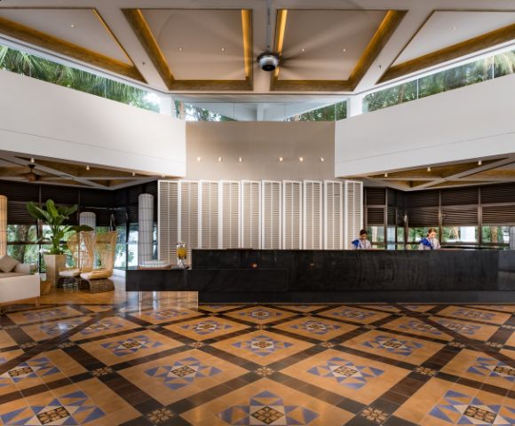 Spacious hotel lobby with a geometric patterned floor, dark reception desk, and modern decor. Two staff members are behind the desk, and lush greenery is visible outside.