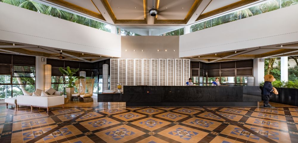 Spacious hotel lobby with a geometric patterned floor, dark reception desk, and modern decor. Two staff members are behind the desk, and lush greenery is visible outside.