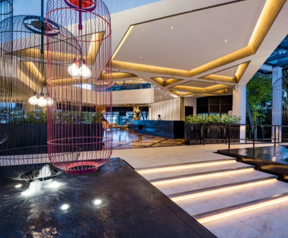 Spacious modern hotel lobby with geometric lighting, two large birdcage light fixtures, sleek white marble steps, and greenery, exuding elegance and warmth.